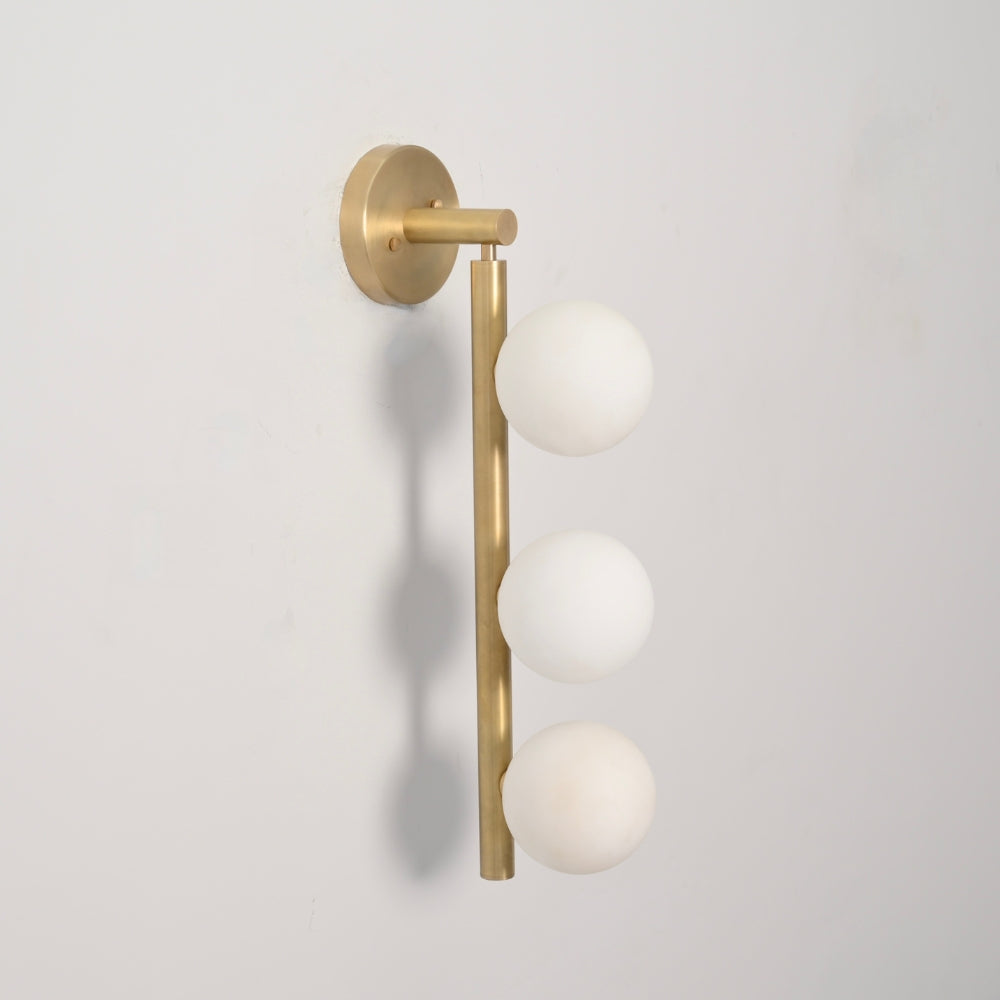 Wall-mounted light fixture with raw brass finish and three white glass globe on a gray background