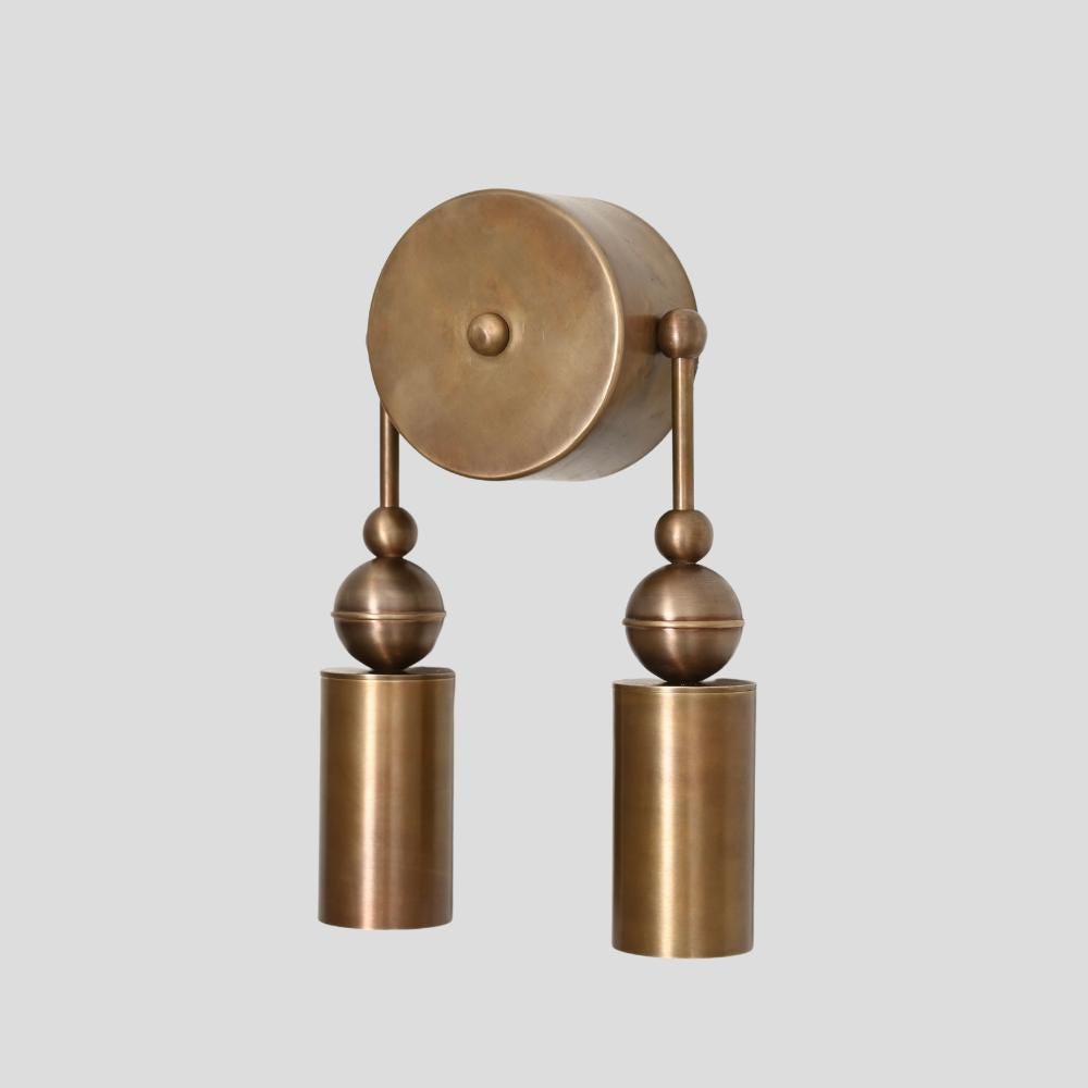 A brass wall sconce with a double cylinder design, featuring a central round brass plate with two vertical rods ending in cylindrical light fixtures.