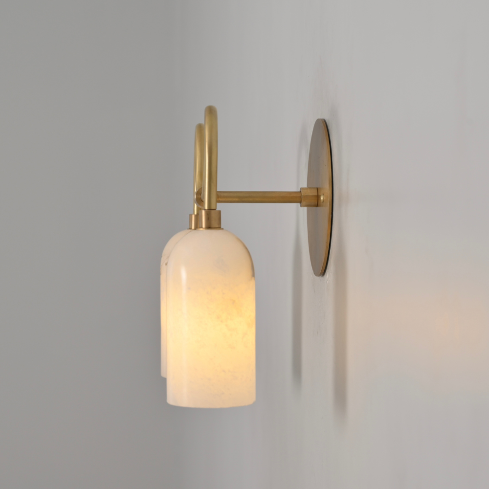 Wall-mounted light fixture with gold arm and white glass shade on a gray wall.