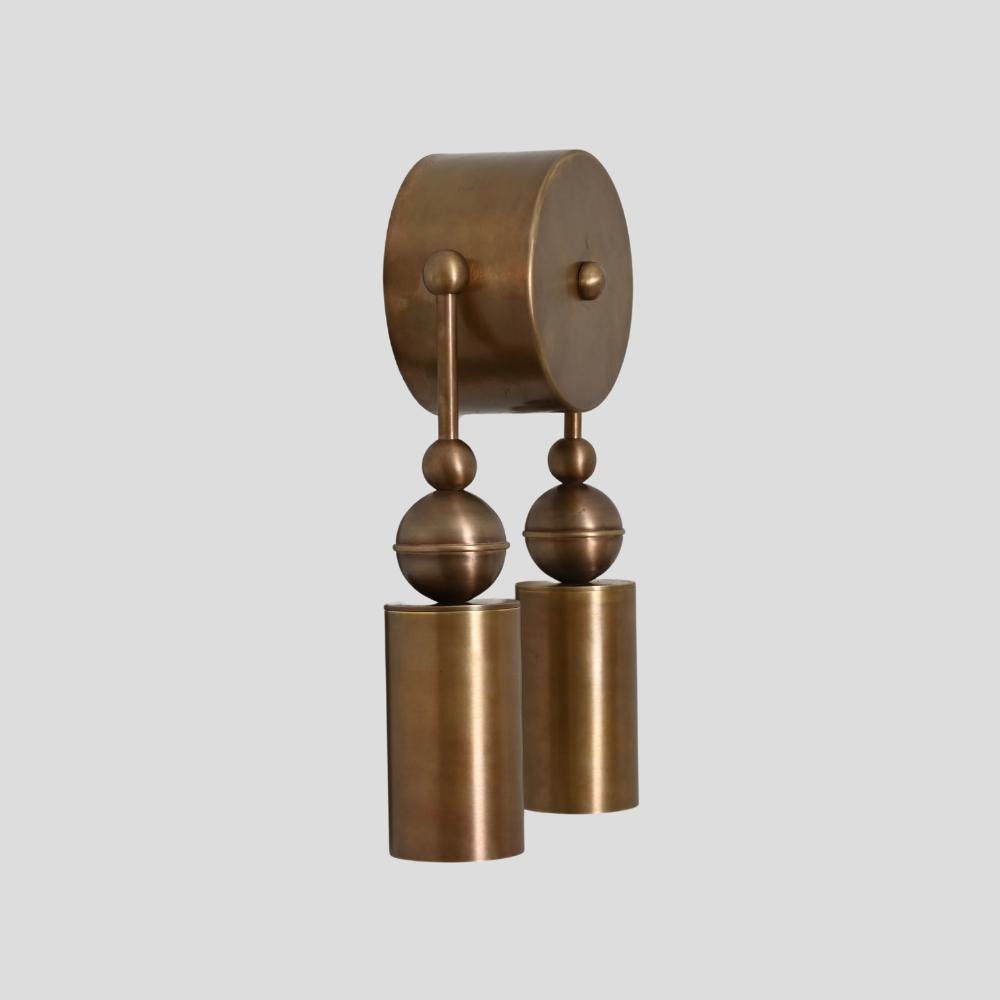 A brass wall sconce with a double cylinder design, featuring a central round brass plate with two vertical rods ending in cylindrical light fixtures.