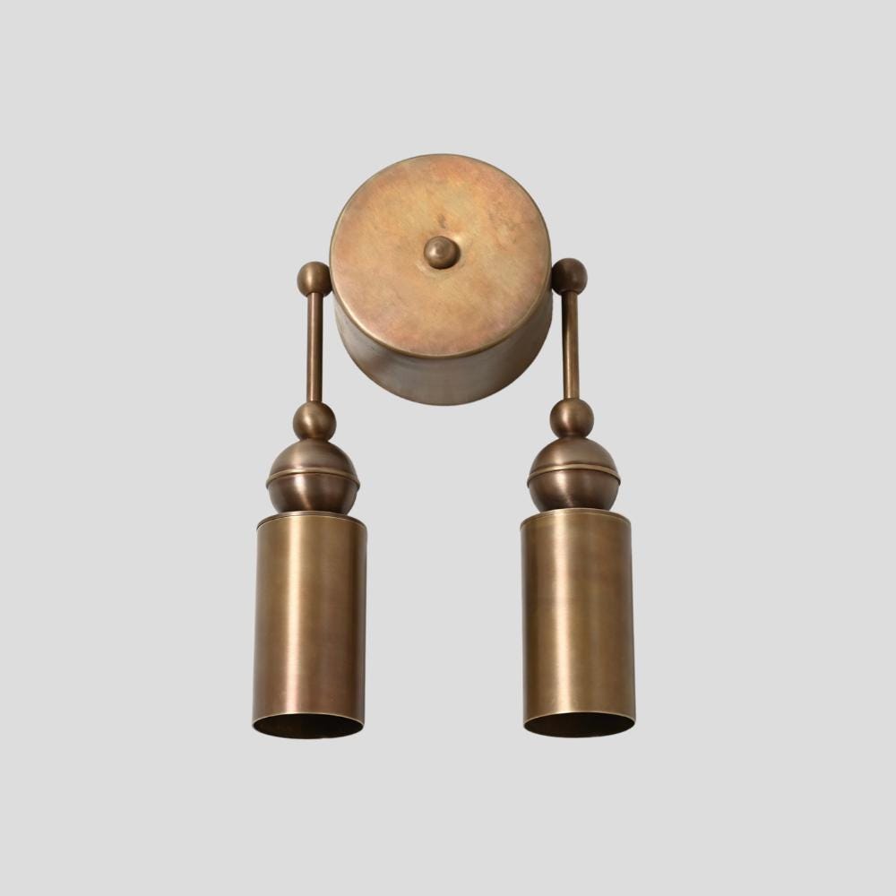 A brass wall sconce with a double cylinder design, featuring a central round brass plate with two vertical rods ending in cylindrical light fixtures.